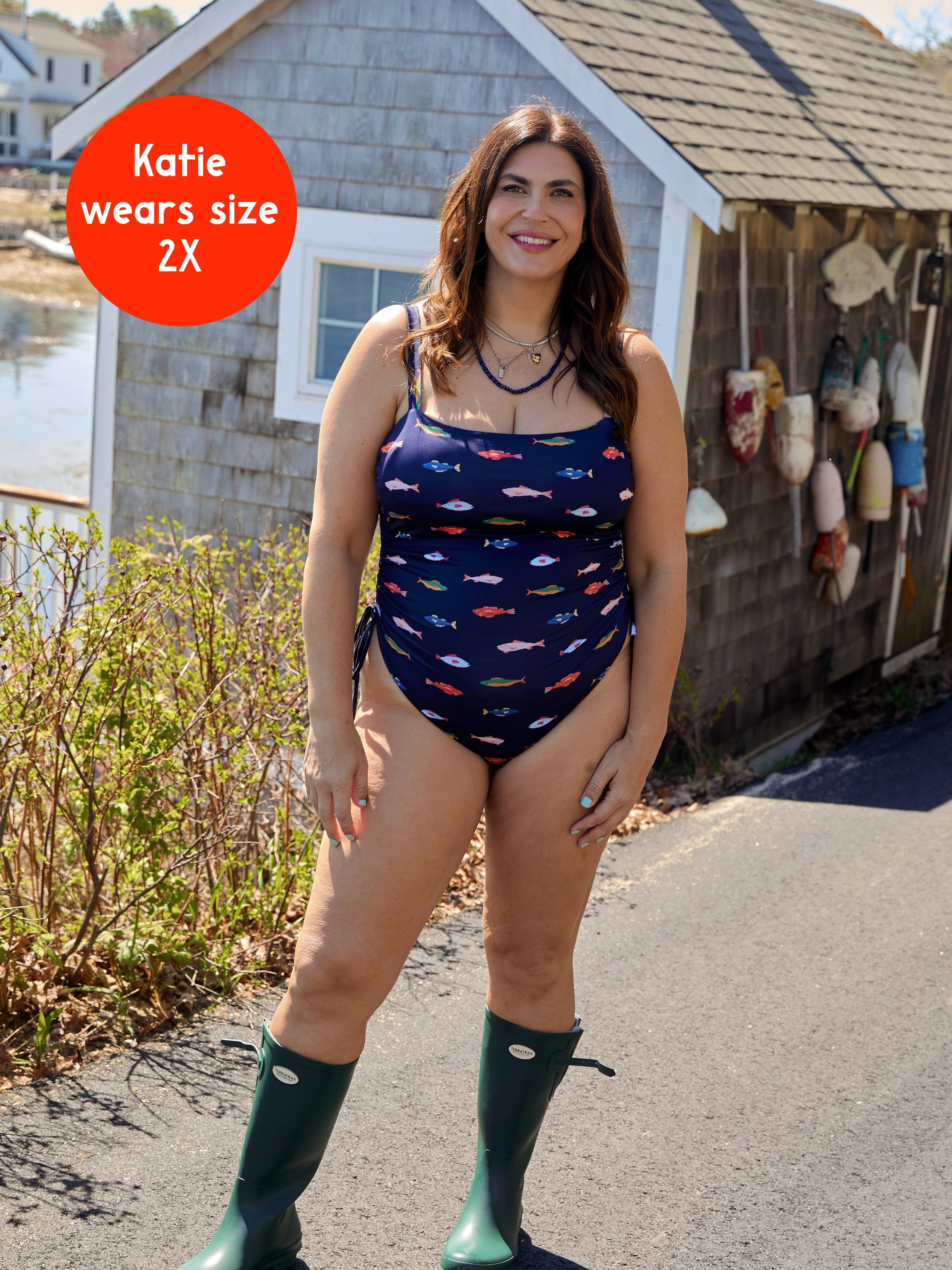 A woman with long brown hair smiles outside in the Kitty and Vibe PREORDER: Ruched Square Neck One Piece in Harbor, featuring adjustable straps, with green boots. “Katie wears size 2X” appears in a red circle. A wooden building and waterfront are behind her.