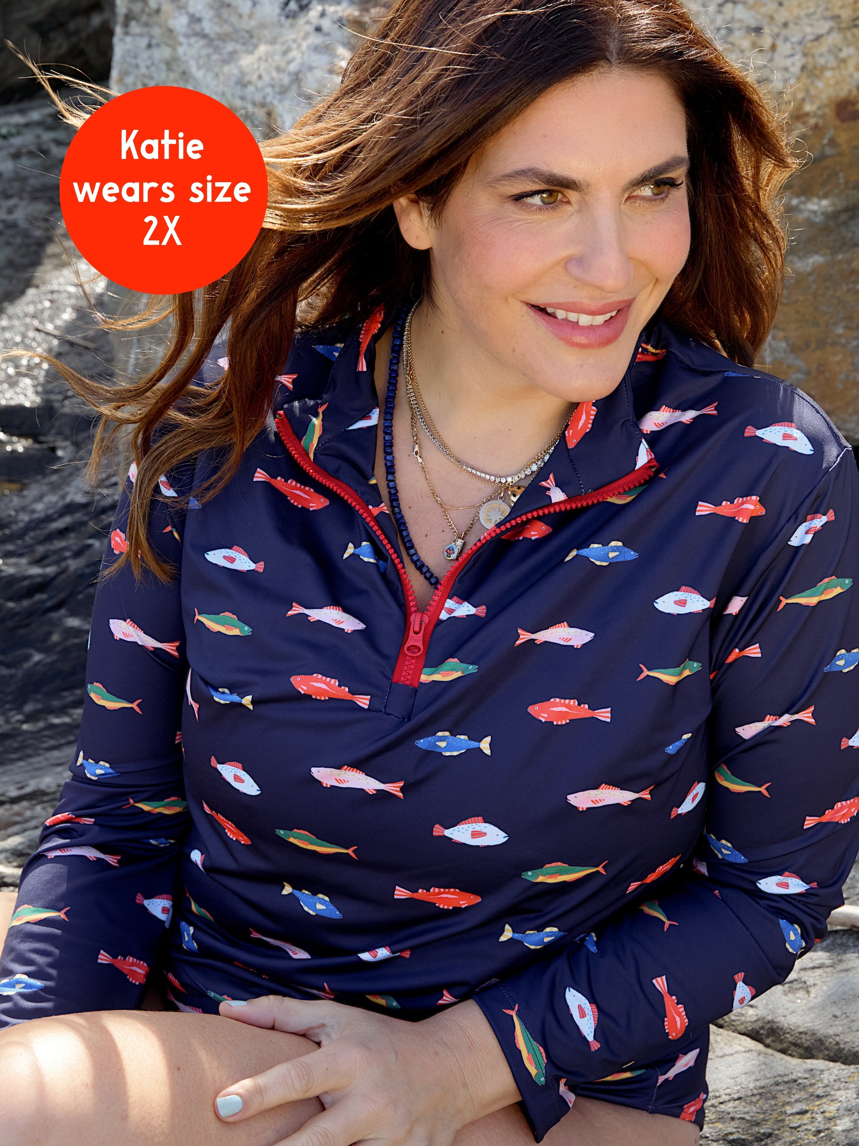 A woman with wavy brown hair smiles outdoors on rocks, wearing the Kitty and Vibe PREORDER: Long Sleeve Rashguard in Harbor (SPF 50+). A red circle reads: Katie wears size 2X.