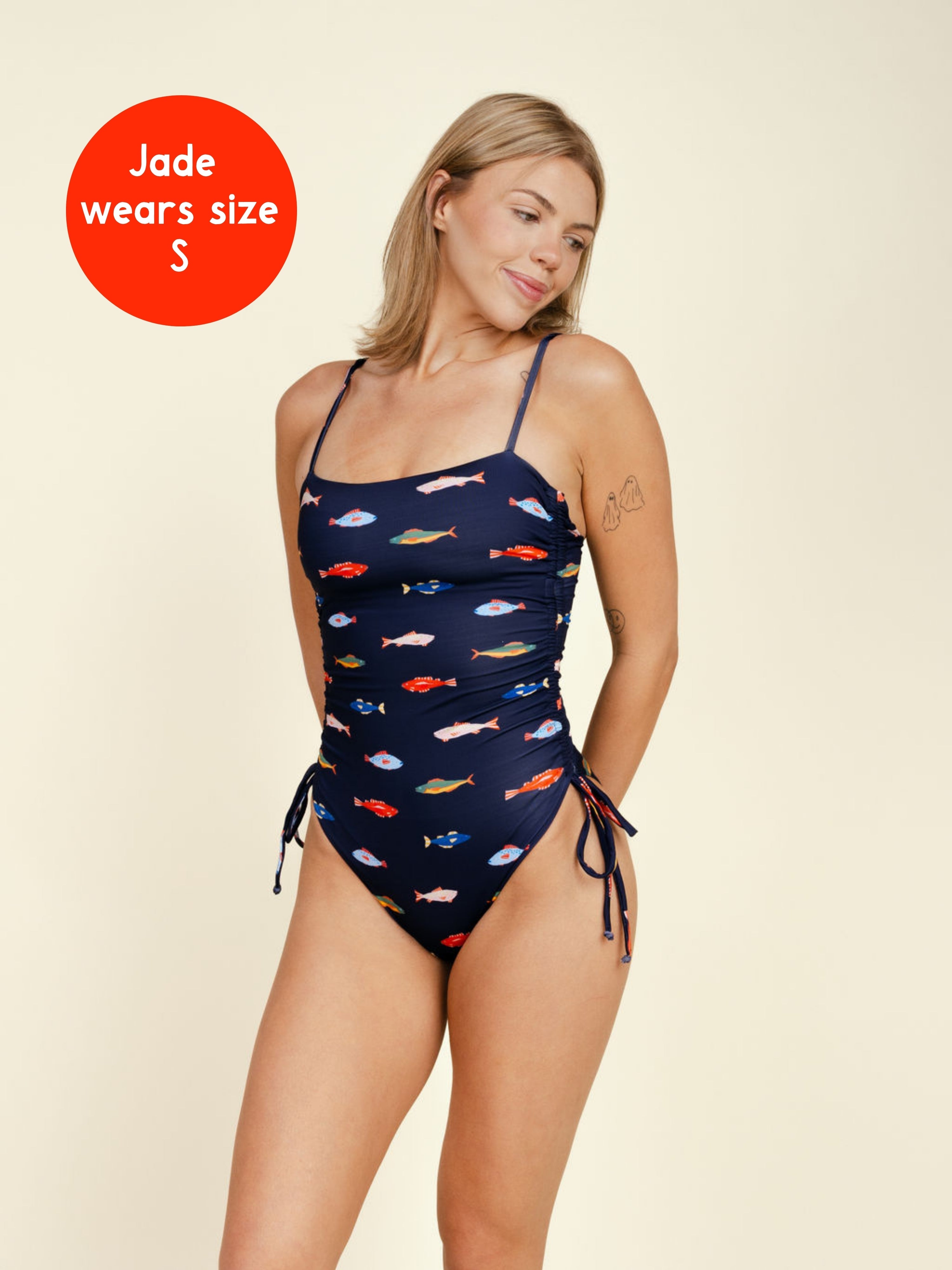 A smiling blonde woman wears the Kitty and Vibe PREORDER: Ruched Square Neck One Piece in Harbor, a navy swimsuit with a colorful fish print, side ties, and adjustable straps. A red circle says, "Jade wears size S.