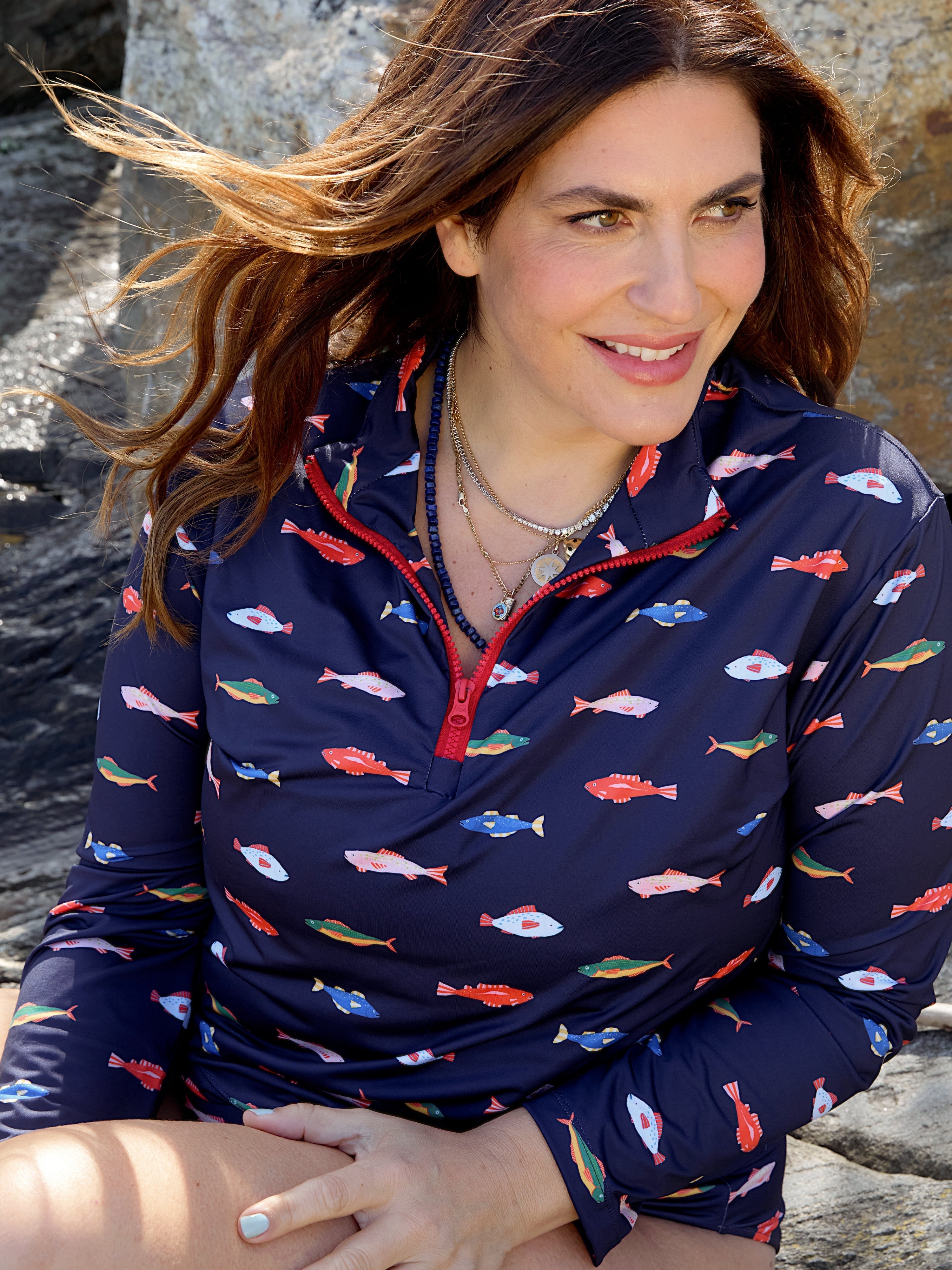 A woman with long brown hair sits on rocks, smiling off-camera. She wears the Kitty and Vibe Long Sleeve Rashguard in Harbor with a colorful fish pattern and zip-front closure. Sunlight highlights her hair and face.