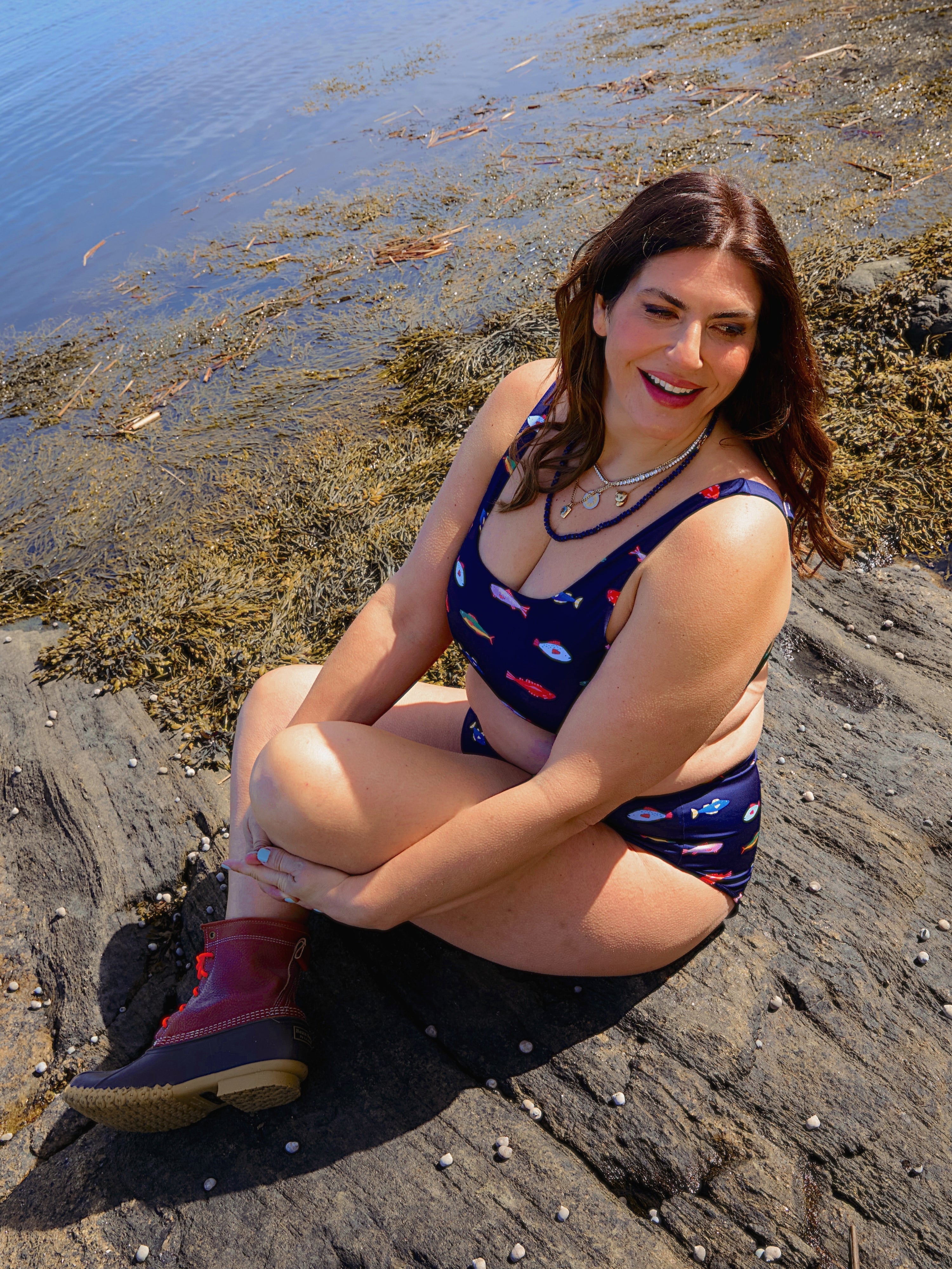 A woman wears the Kitty and Vibe Scoop Top in Harbor bikini, featuring adjustable straps, as she sits smiling on a rock by the water, surrounded by seaweed and shells on a sunny day.