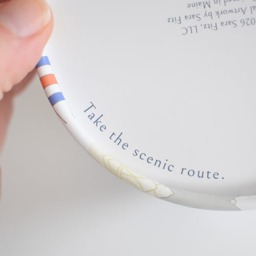 A hand holds a circular piece from the Sara Fitz New England Puzzle, with the text Take the scenic route printed along the inner edge of the 500-piece design.