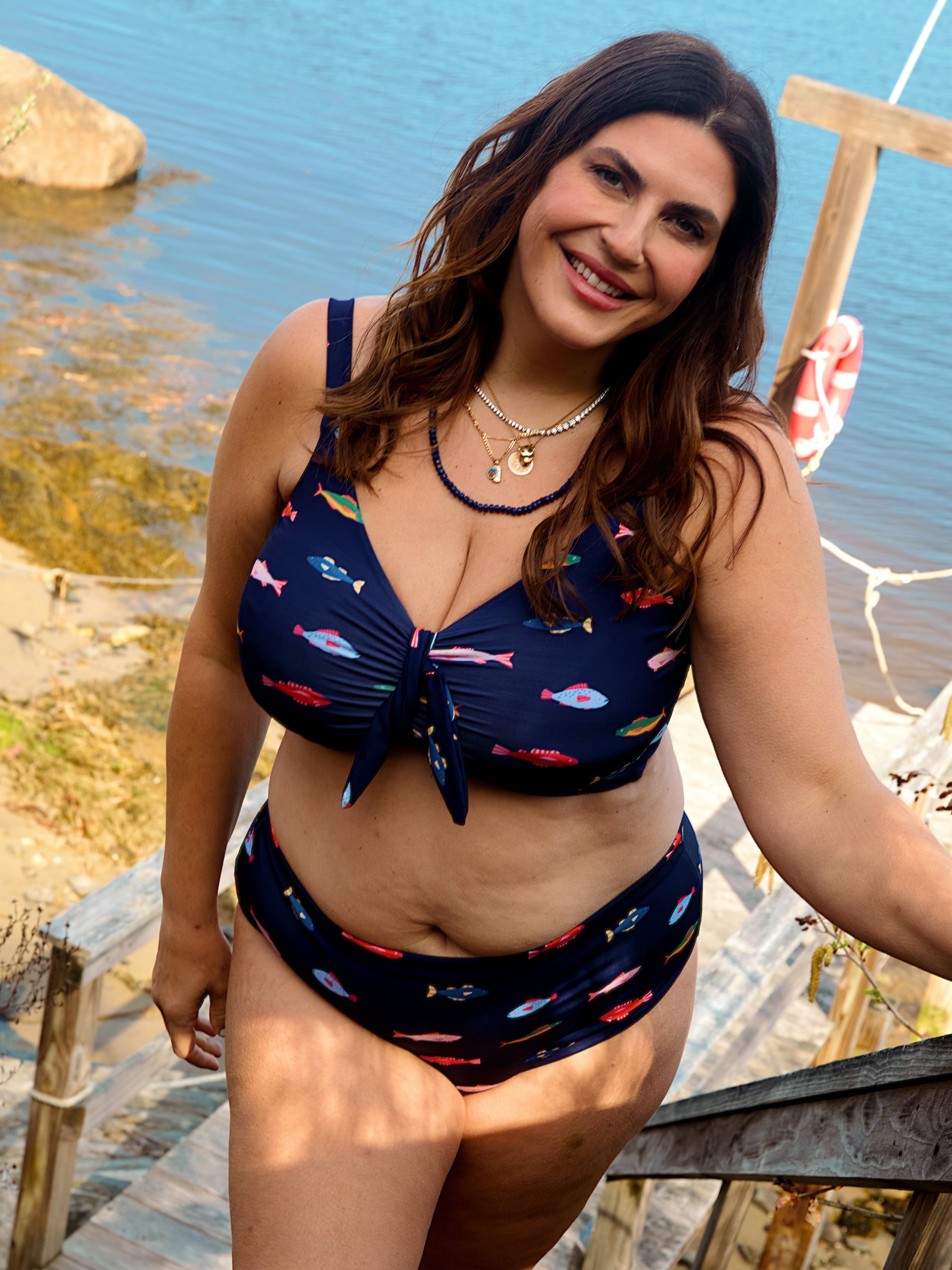A smiling woman with long brown hair wears the Kitty and Vibe Front Tie Top in Harbor with adjustable straps, standing on wooden steps by the lakeshore with water and rocks in the background.