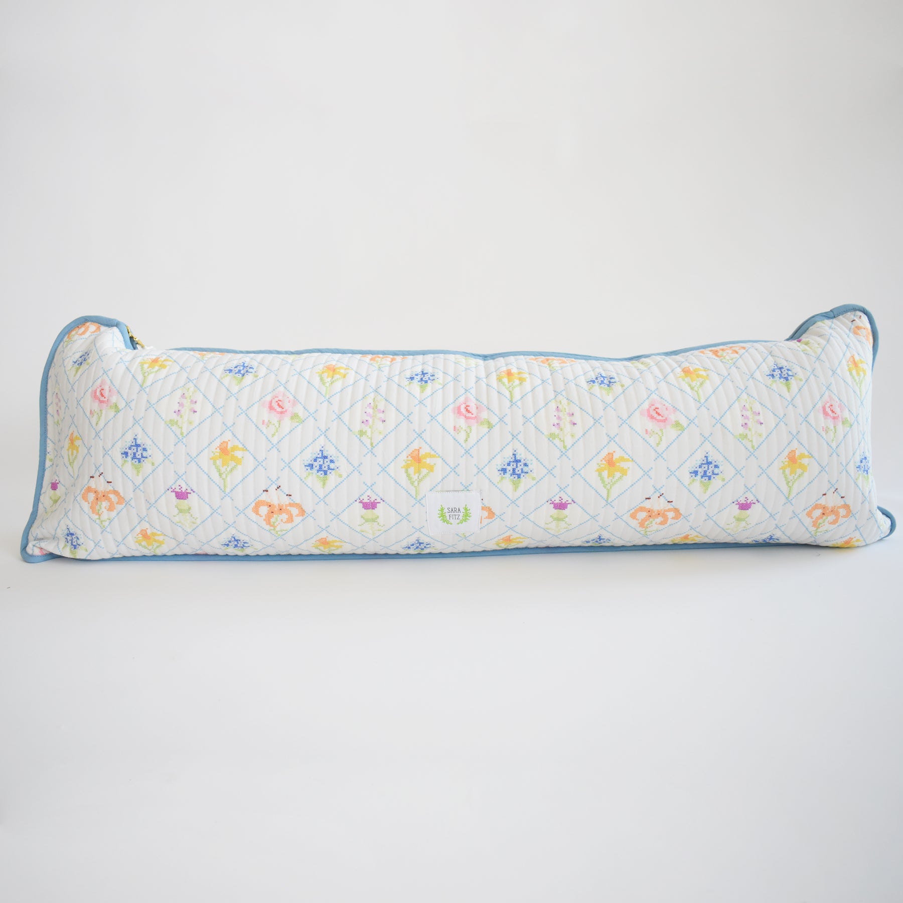 The Sara Fitz Lumbar Pillow, Cross Stitch Floral, is a long, rectangular cotton quilted pillow made in India with a white background, blue edging, and a grid of colorful floral and small motif designs.