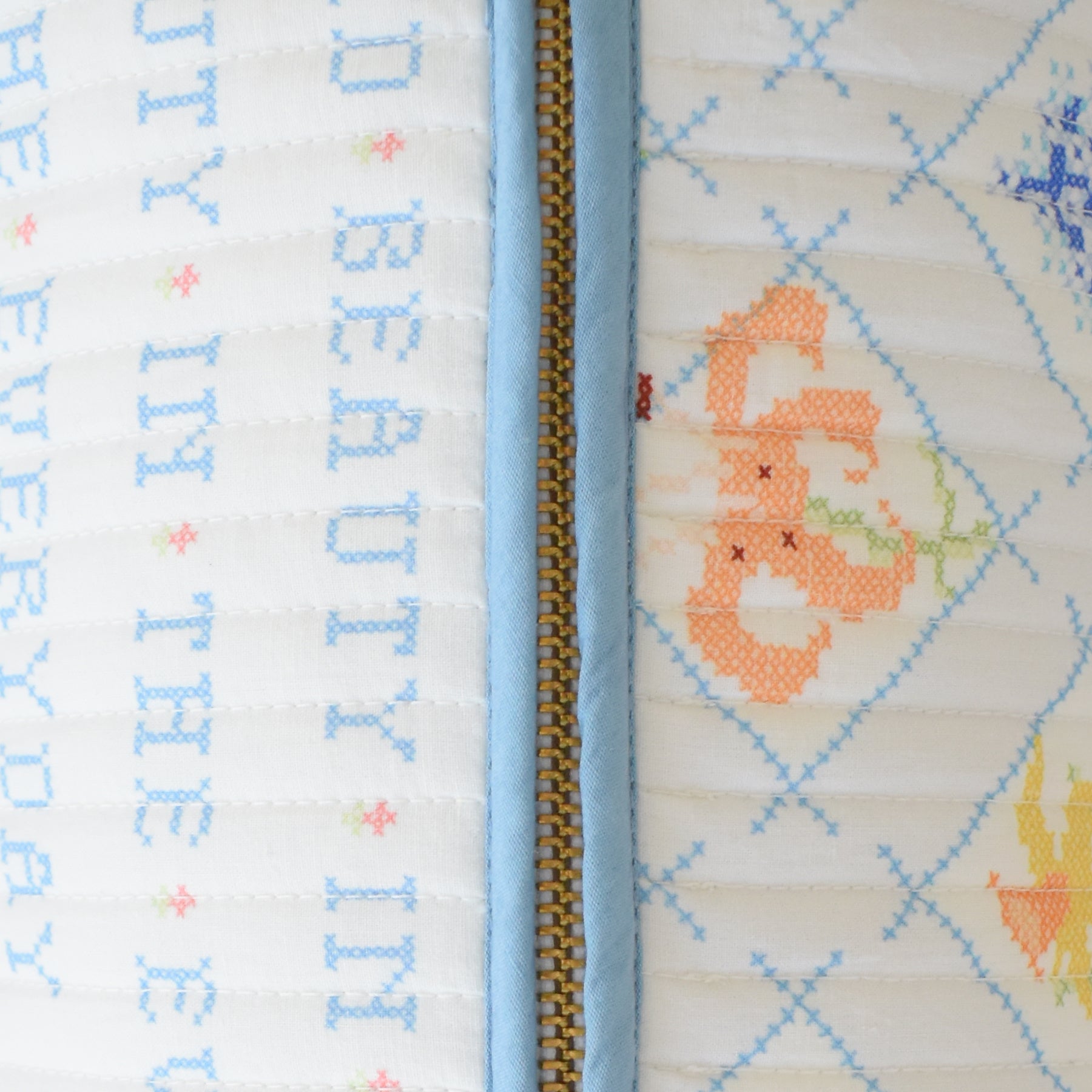 Close-up of the Sara Fitz Lumbar Pillow, Cross Stitch Floral: a cotton quilted pillow with a blue zipper, pastel floral cross-stitched designs, blue text on white fabric. Made in India.