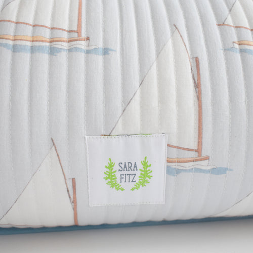 A close-up of the Sara Fitz Lumbar Pillow, Sailboat, featuring a soft cotton quilted cover with a sailboat pattern, a white fabric tag with "Sara Fitz" in green and leaf detail, and a plush down alternative insert for comfort.