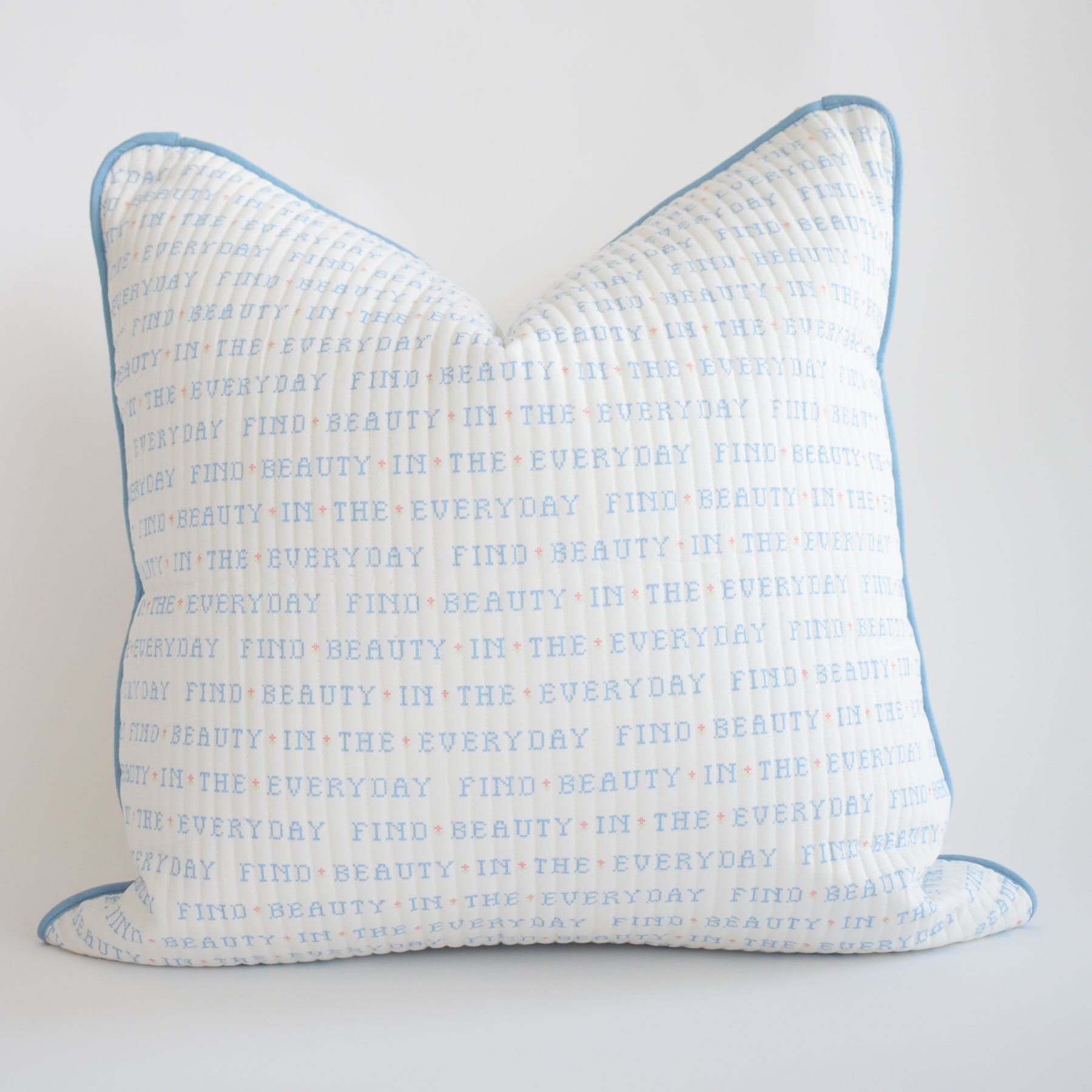 The Sara Fitz Oversized Pillow, Cross Stitch Floral, is a white cotton quilted pillow with blue piping, featuring repeated blue and orange text “FIND BEAUTY IN THE EVERYDAY.” Includes down alternative insert. Made in India.