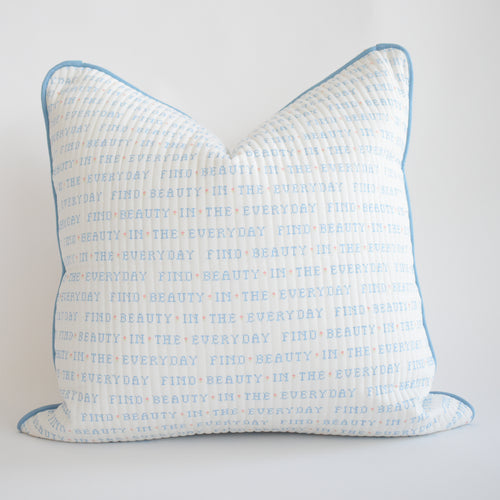 The Sara Fitz Oversized Pillow, Cross Stitch Floral, is a white cotton quilted pillow with blue piping, featuring repeated blue and orange text “FIND BEAUTY IN THE EVERYDAY.” Includes down alternative insert. Made in India.