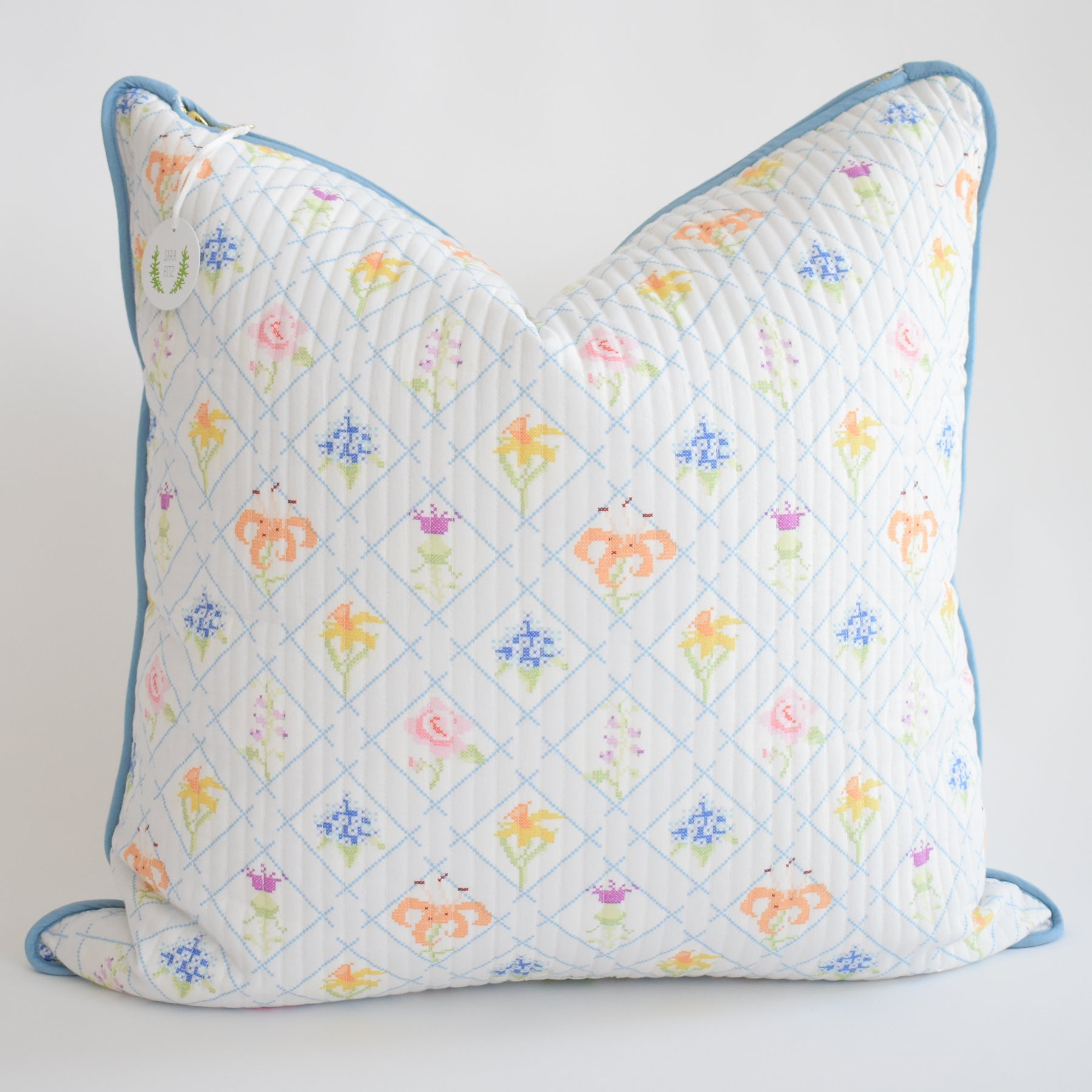 The Sara Fitz Oversized Pillow, Cross Stitch Floral, features a white cotton quilted cover with pastel floral and diamond patterns, light blue piping, a small round tag, down alternative insert, and is made in India.