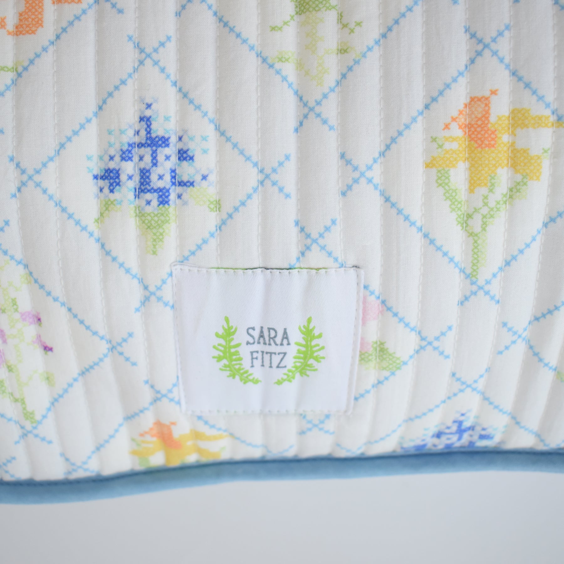 A close-up of the Sara Fitz Oversized Pillow, Cross Stitch Floral, shows a cotton quilted cover with blue diamonds and colorful floral embroidery. A central white label reads SARA FITZ, surrounded by a green leafy wreath. Down alternative insert; made in India.
