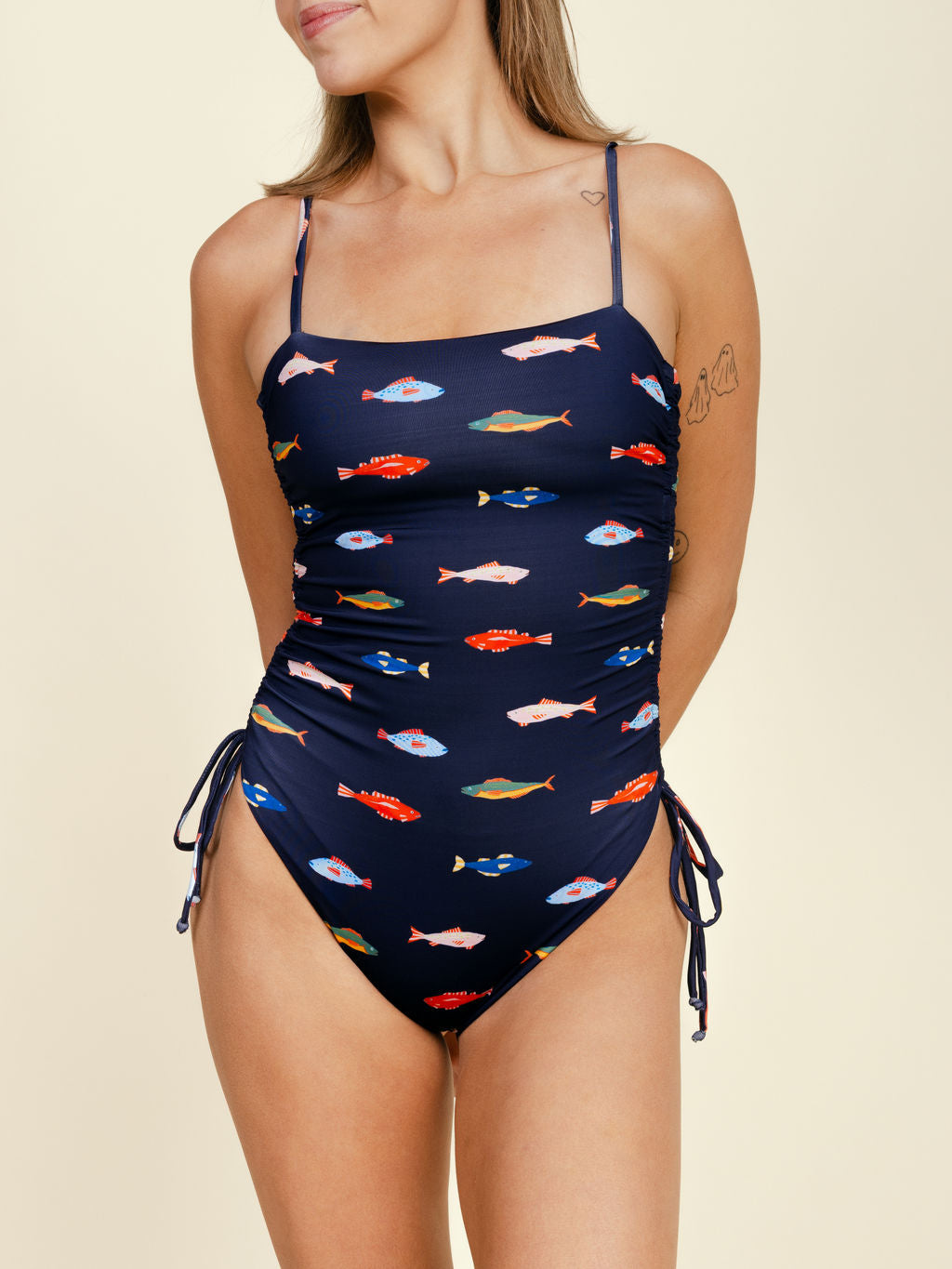A woman models the Kitty and Vibe Ruched Square Neck One Piece in Harbor—featuring colorful fish print, thin adjustable straps, a built-in shelf bra, and side drawstrings—against a neutral background.