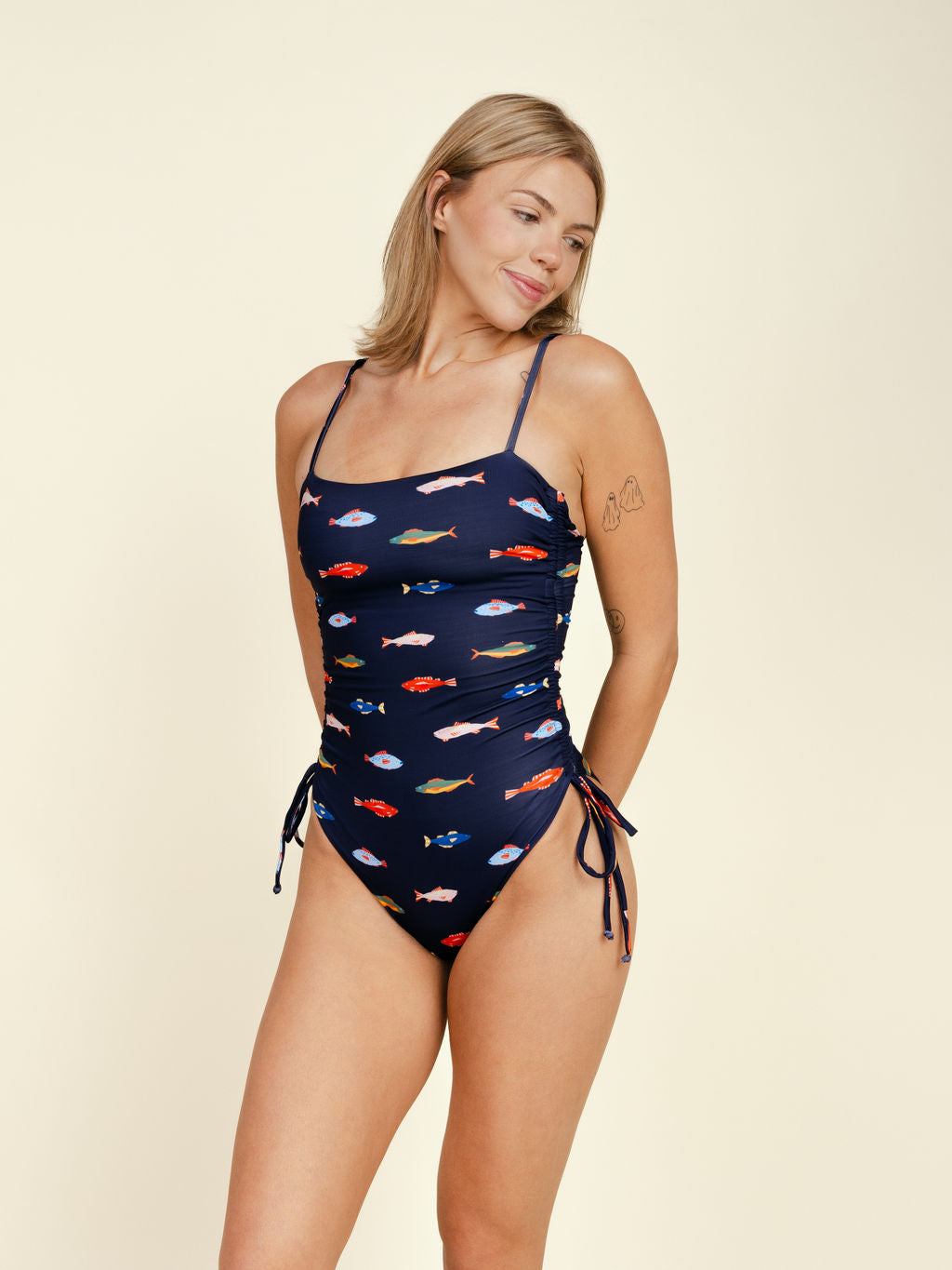 A blonde woman wears the Kitty and Vibe Ruched Square Neck One Piece in Harbor, featuring colorful fish print, side ties, and adjustable straps. She's standing and smiling with her arms behind her back against a plain light background.