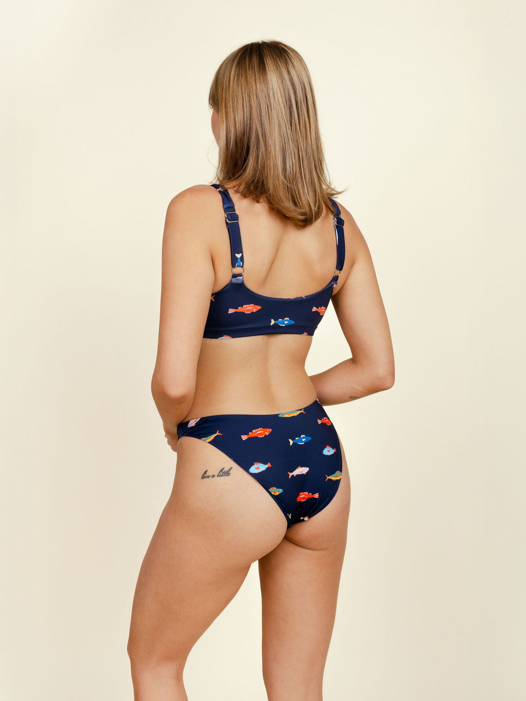 A woman with shoulder-length blonde hair stands facing away, wearing Kitty and Vibe's Brief Bottoms in Harbor—a navy blue, mid-rise bikini with a colorful fish pattern. She has a small upper left thigh tattoo and poses against a plain background.