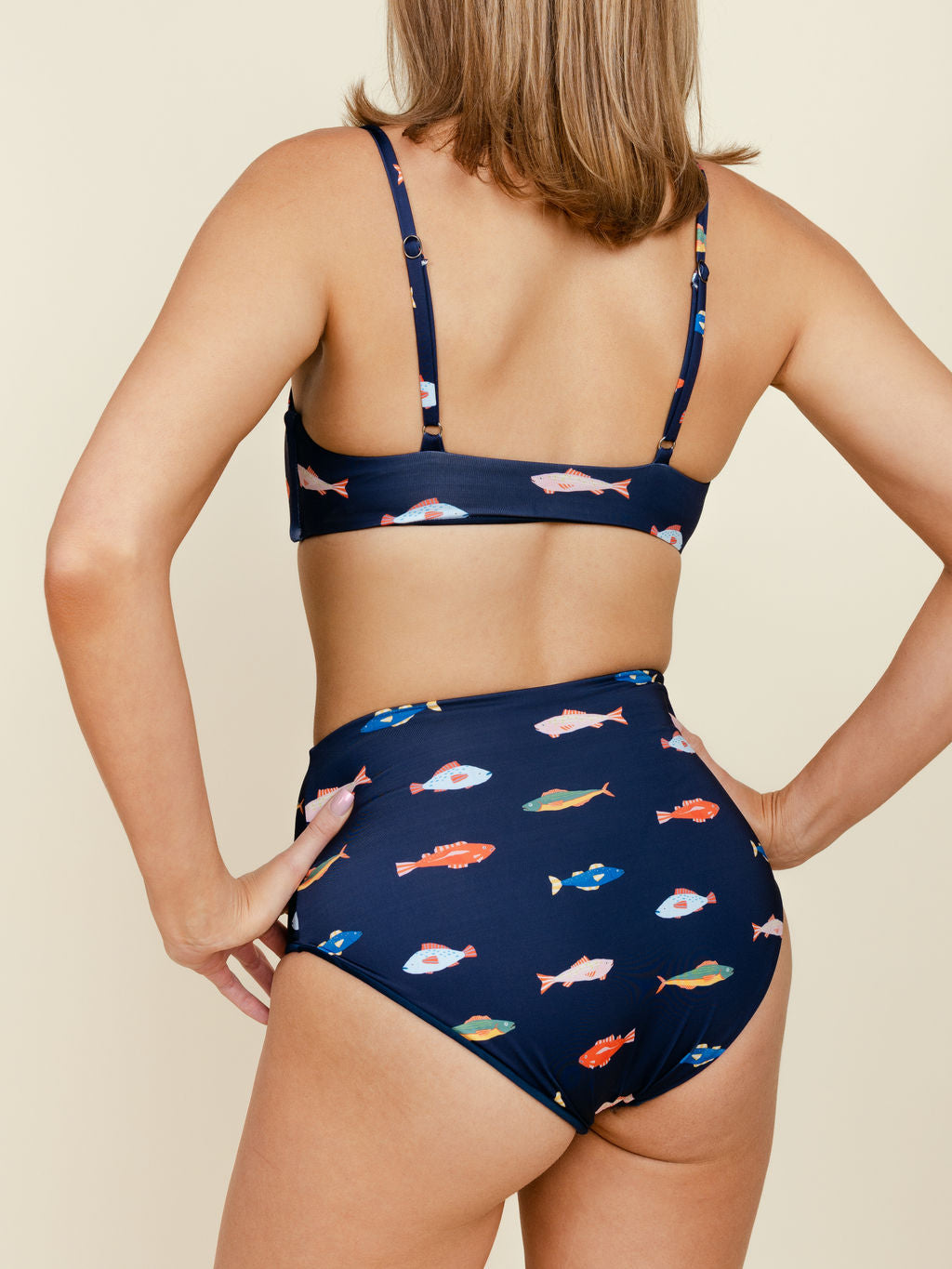 A person stands with hands on hips, modeling the Kitty and Vibe High Waisted Full Coverage Bottoms in Harbor, featuring a colorful fish pattern. The back view is shown against a plain beige background.