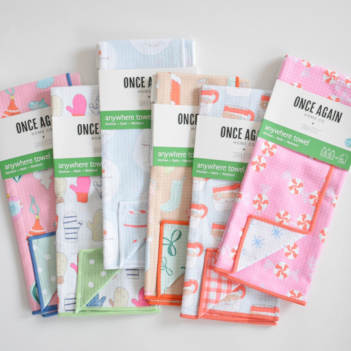 Five folded, absorbent Sara Fitz Anywhere Towel, Peppermint towels with winter-themed patterns including mittens, candy canes, and snowflakes, each secured with a branded paper sleeve.