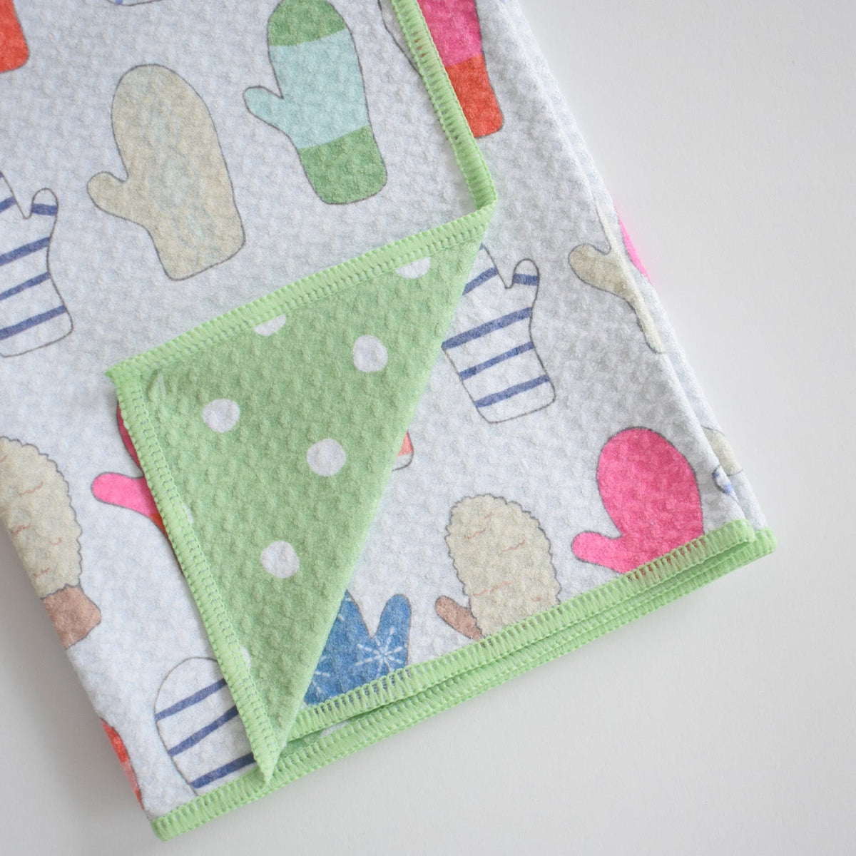 Light gray Sara Fitz Anywhere Towel, Mittens, patterned with colorful mittens and folded to reveal an absorbent green interior with white polka dots and stitched edges.