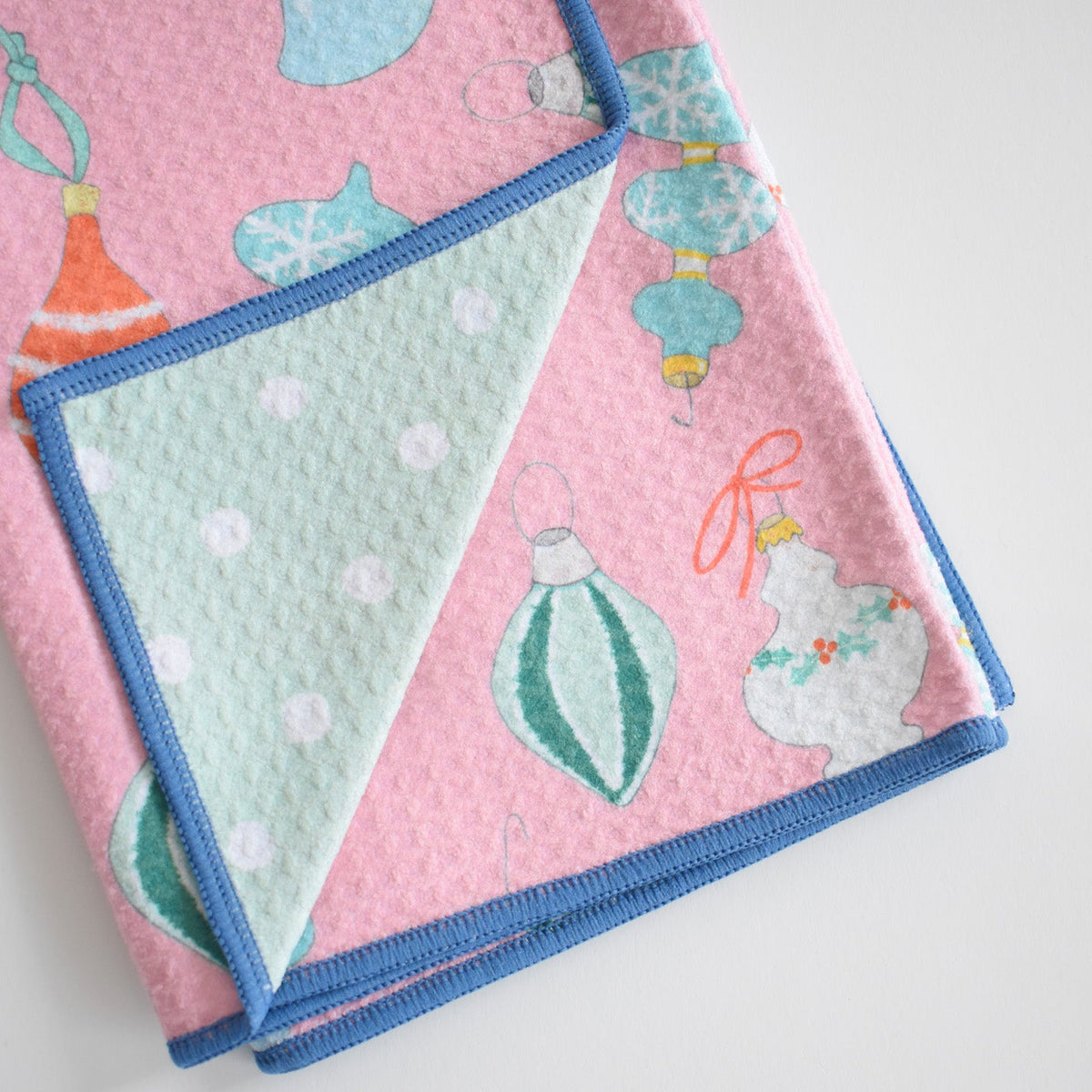 Sara Fitz Anywhere Towel, Ornament: a pink absorbent towel with a pattern of Christmas ornaments, folded to reveal a light green underside with white polka dots and a blue stitched edge.