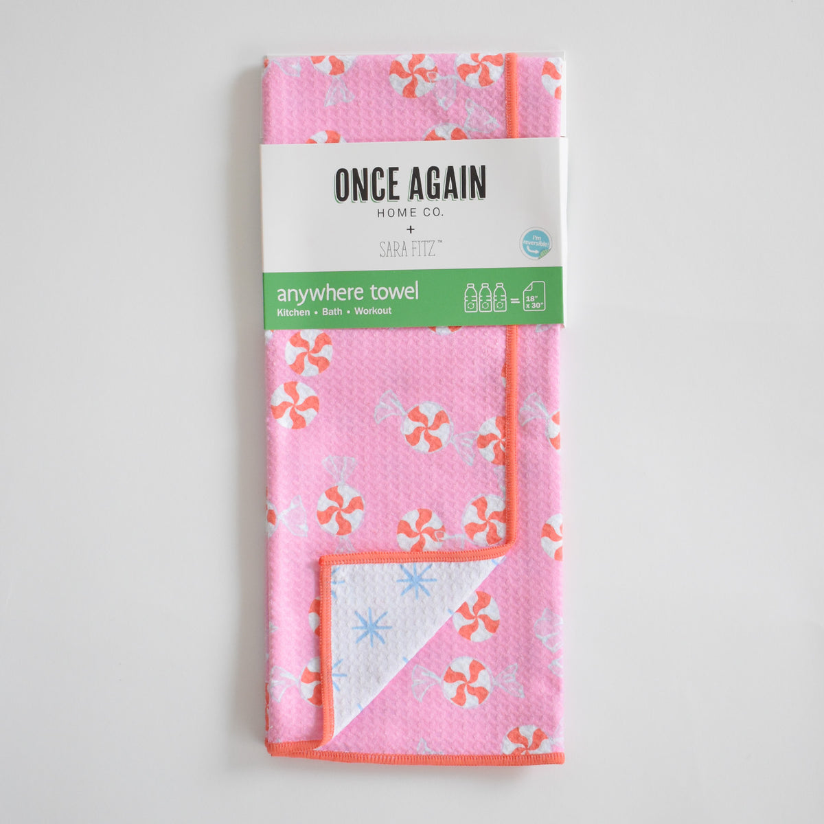 Sara Fitz Anywhere Towel, Peppermint, featuring a pink repeating peppermint candy pattern with an orange trim, wrapped in a Once Again Home Co. label and made of absorbent fabric.