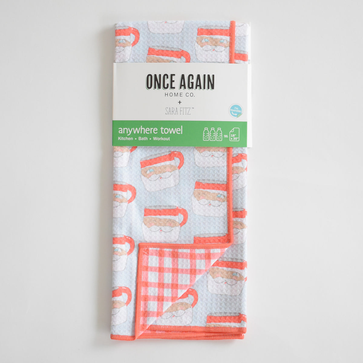A folded Sara Fitz Anywhere Towel, Santa Mug, with a light blue Santa mug print, a coral-colored gingham underside, and a recycled product label.