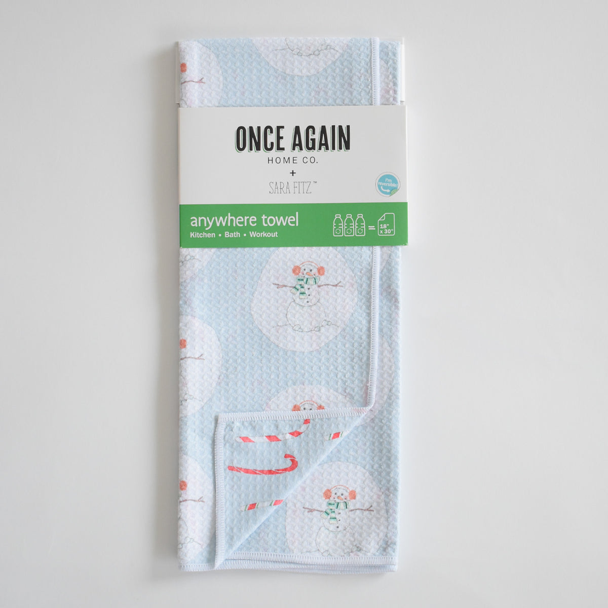 Sara Fitz Anywhere Towel, Happy Snowman, featuring light blue microfiber fabric with a pattern of snowmen in earmuffs and scarves, folded with a branded paper sleeve around the center.