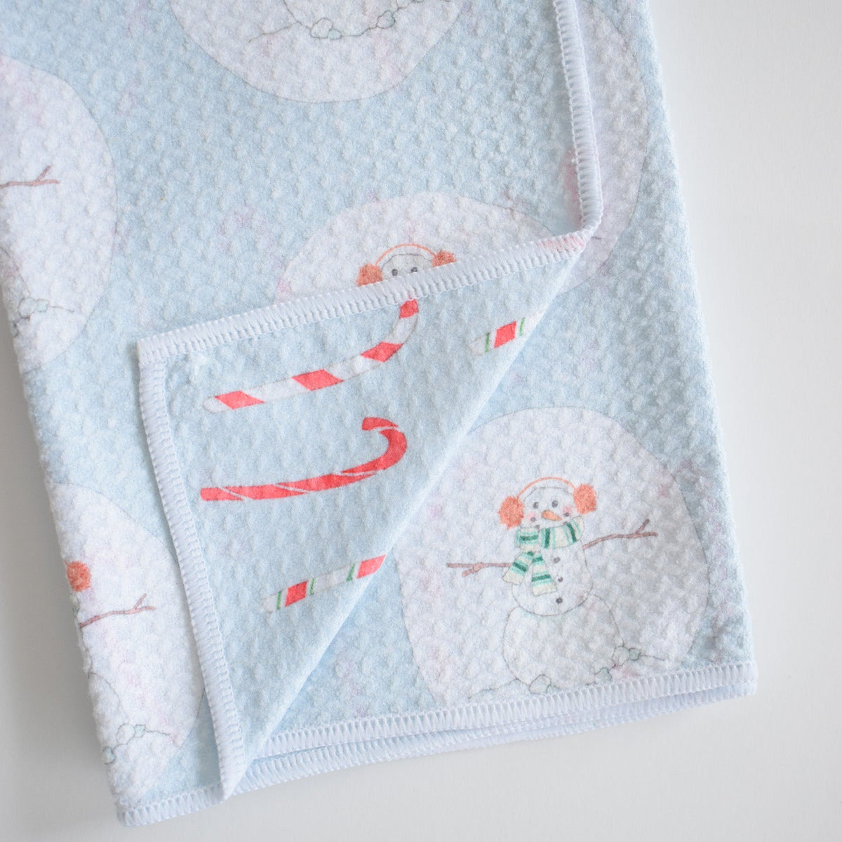 Light blue textured Sara Fitz Anywhere Towel, Happy Snowman, featuring a pattern of snowmen with earmuffs and scarves, and red and white candy canes, made from absorbent fabric.