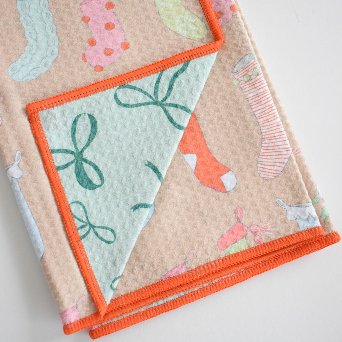 A folded, absorbent tan Anywhere Towel, Stockings featuring a pattern of colorful Christmas stockings, a mint green lining with a bow print, and an orange stitched edge.