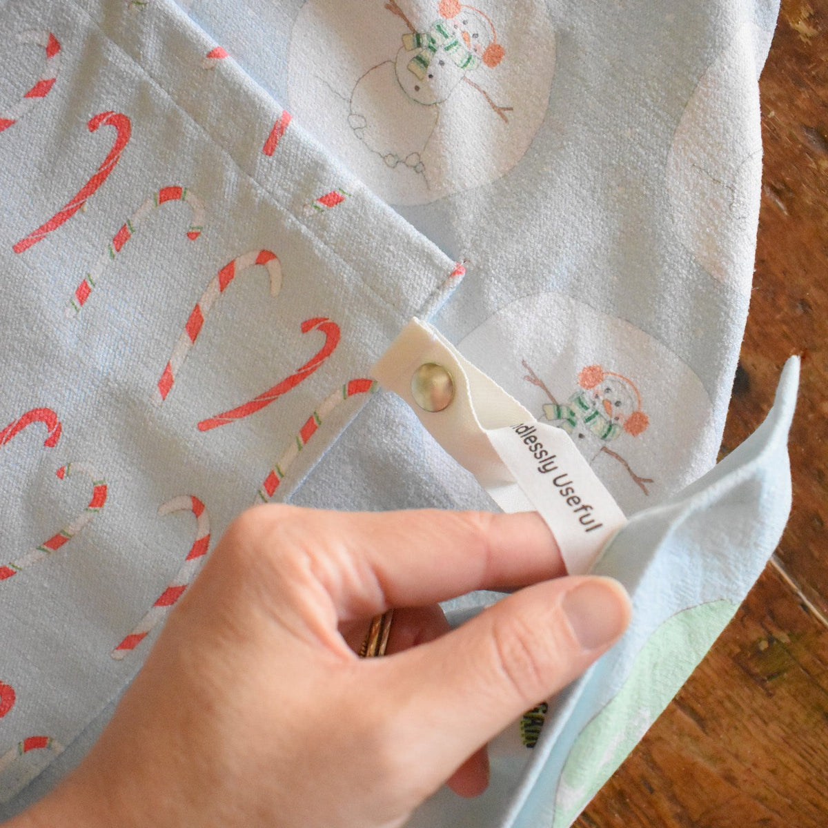 A hand holds a tab on a light blue Attach + Go Apron, Happy Snowman featuring a pattern of snowmen and candy canes, with the text Endlessly Useful printed on the tab.