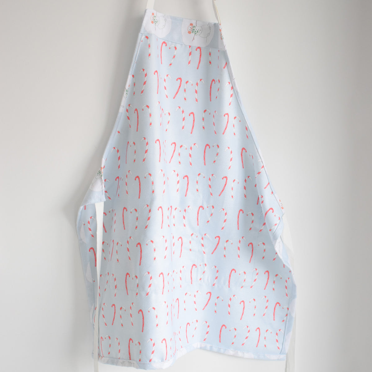 Light blue Attach + Go Apron, Happy Snowman patterned with red and white candy canes hanging against a white wall.
