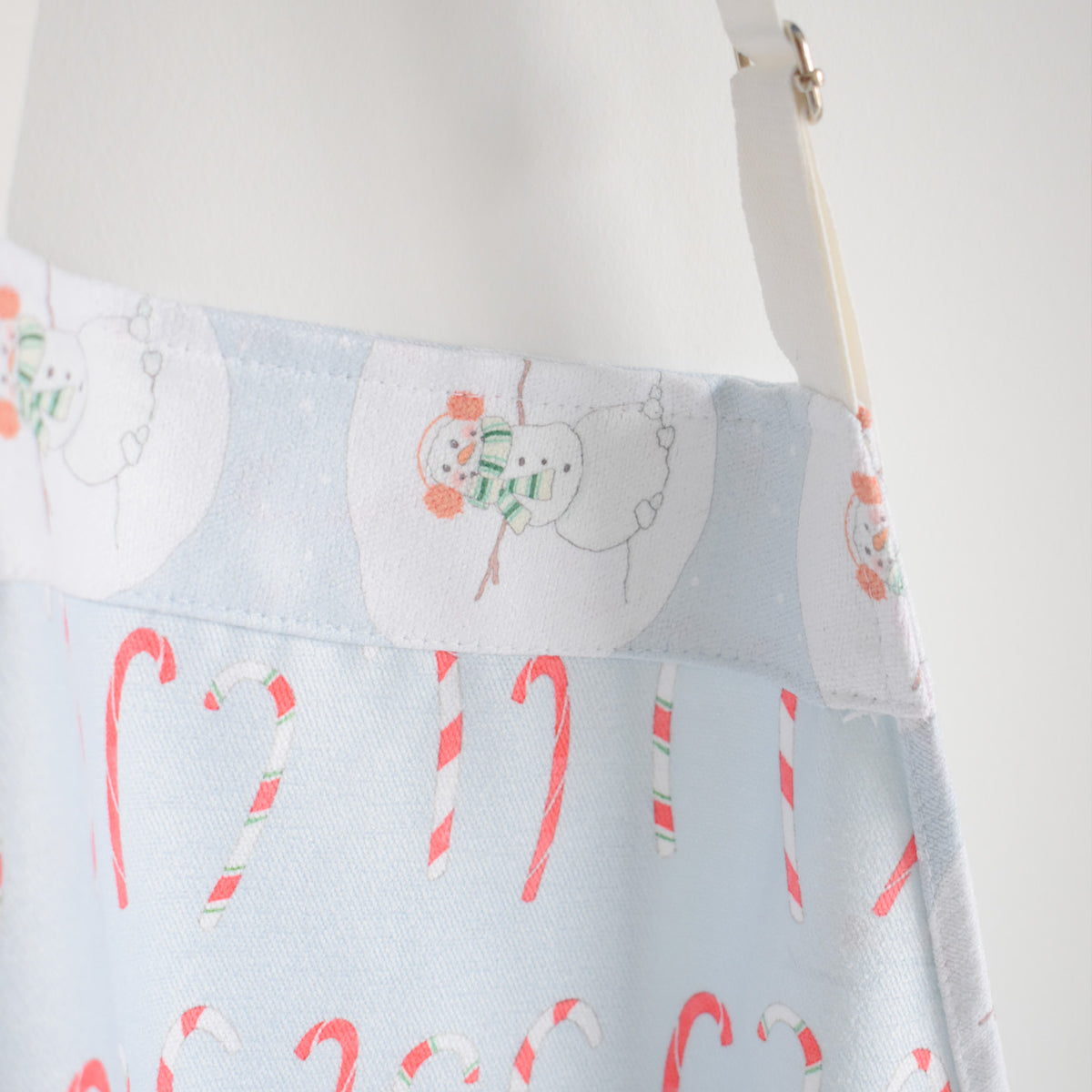 Sara Fitz Attach + Go Apron, Happy Snowman, a light blue apron patterned with red and white candy canes and illustrated snowmen wearing scarves.