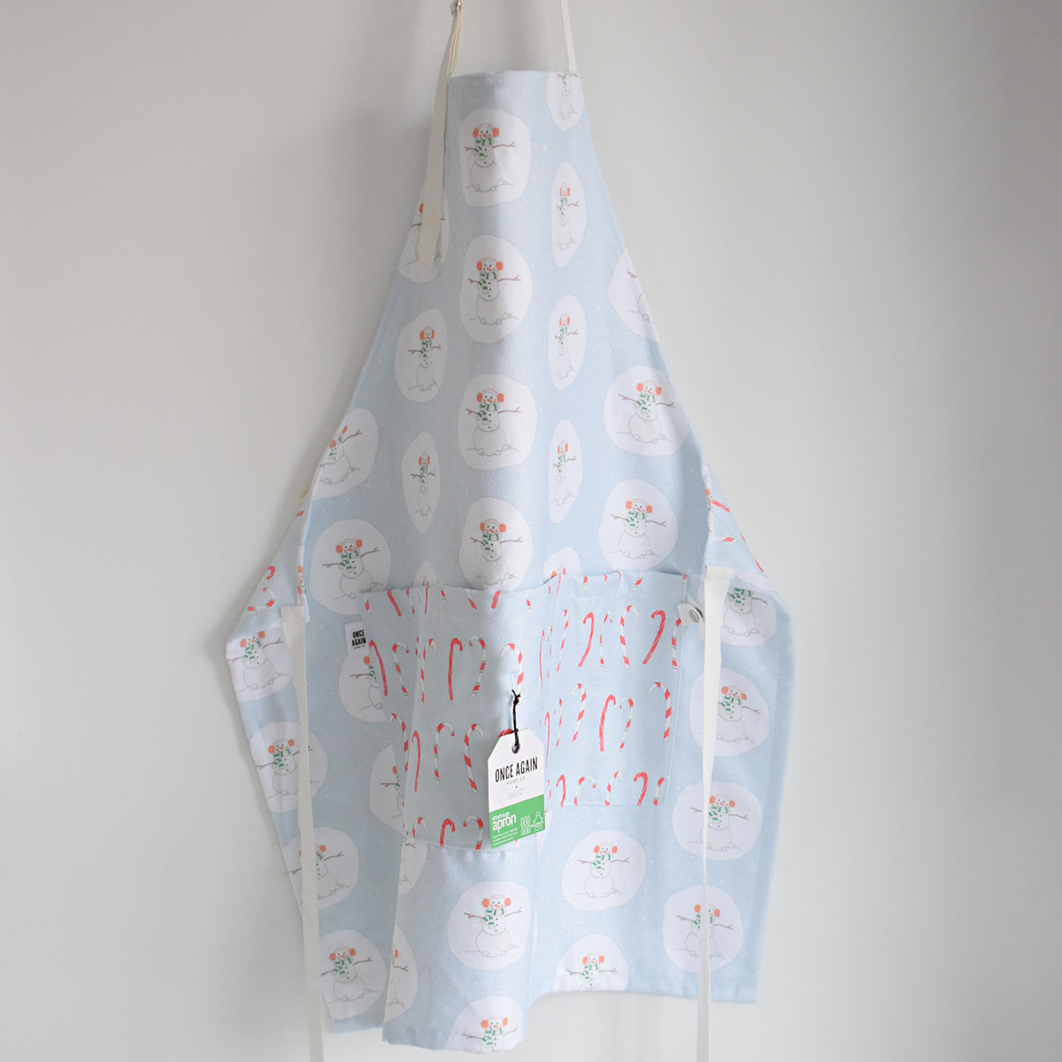 Sara Fitz Attach + Go Apron, Happy Snowman, featuring a light blue pattern of snowmen and candy canes, hanging against a white wall.