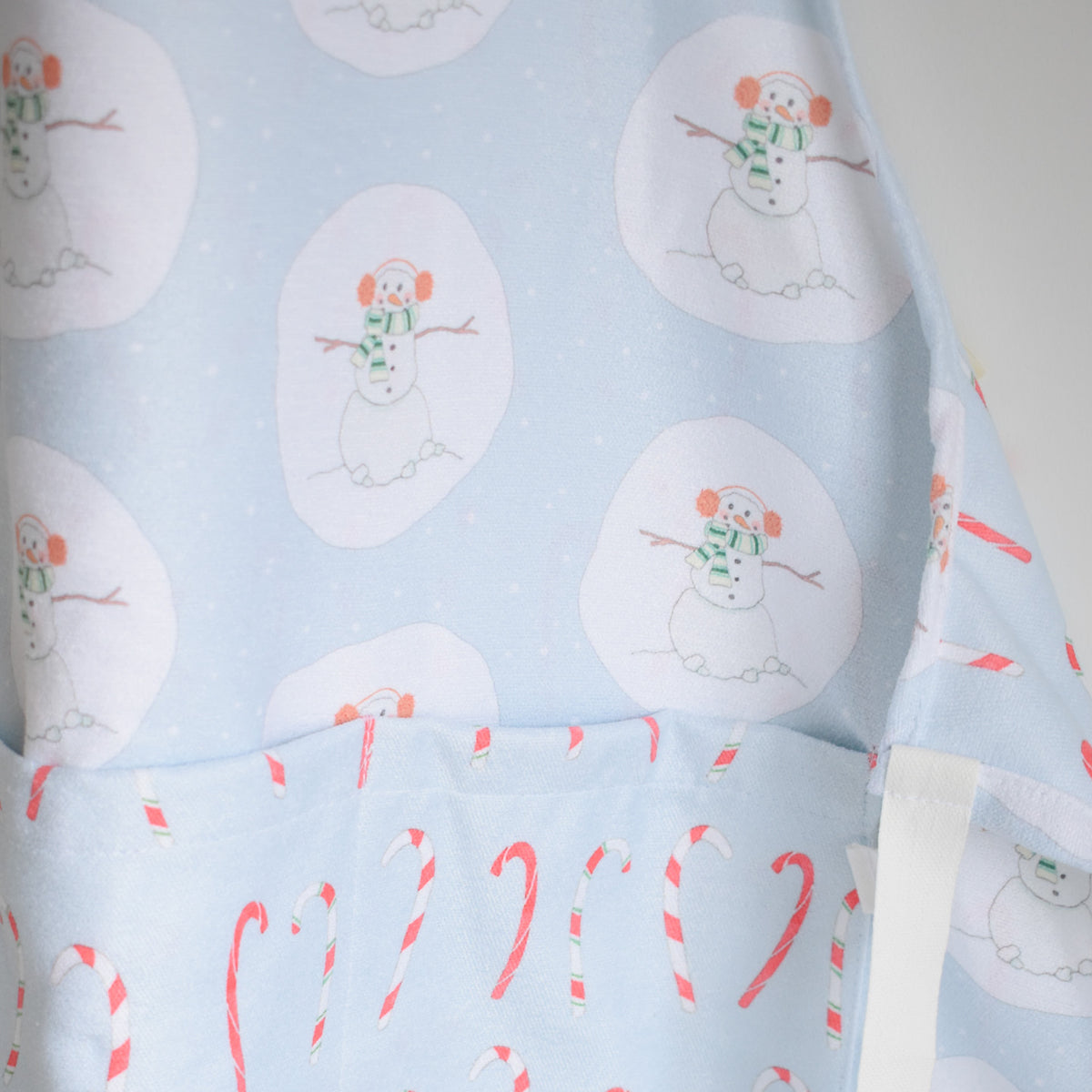 Sara Fitz Attach + Go Apron, Happy Snowman, featuring a light blue pattern of snowmen in earmuffs and a pocket decorated with red and white candy canes.