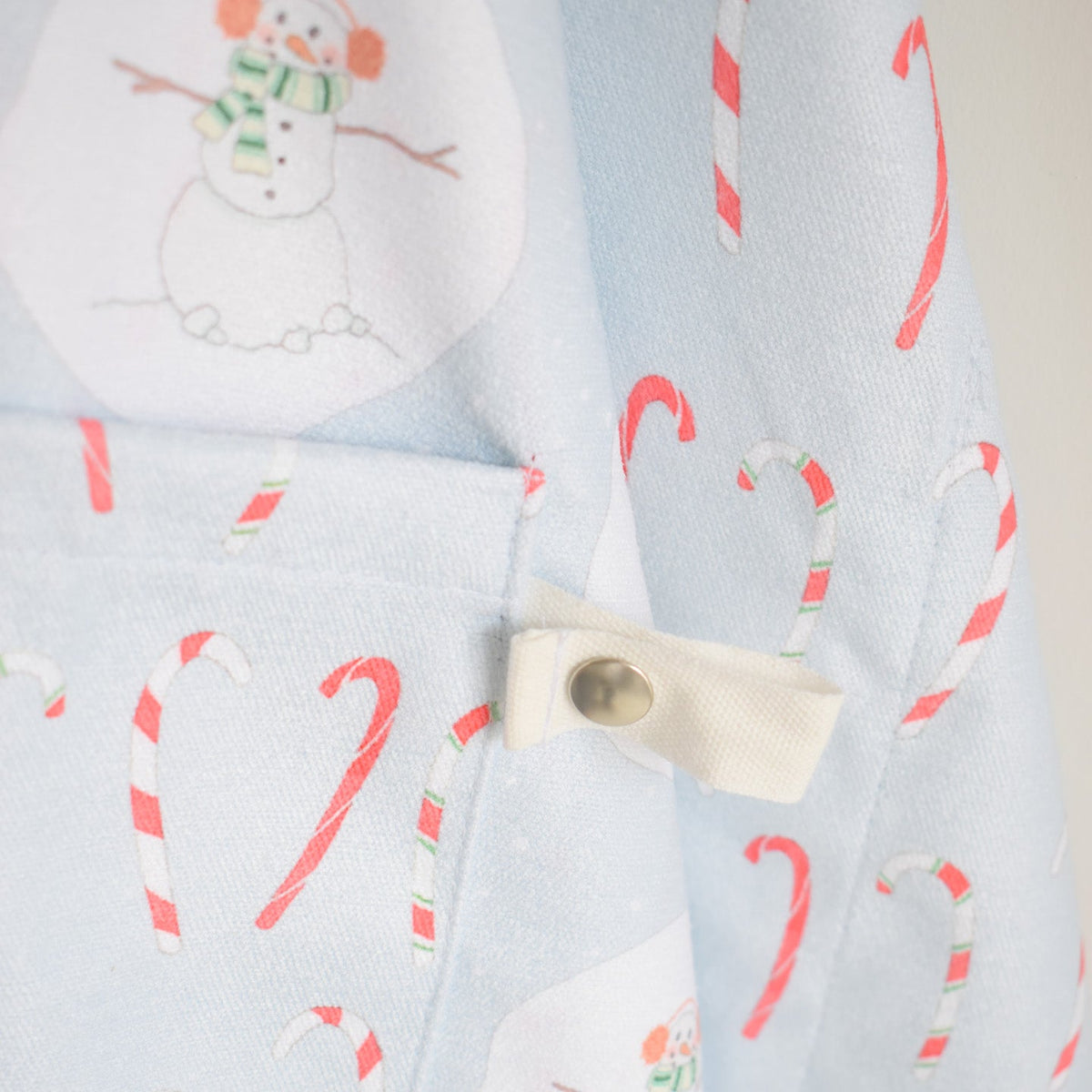 Sara Fitz Attach + Go Apron, Happy Snowman, featuring a light blue fabric with a snowman and candy cane pattern, and a side tab with a metal snap closure.
