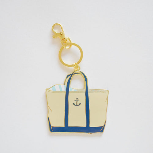 The Canvas Tote Bag Charm by Sara Fitz features a gold keychain with an enamel beige tote bag, blue handles and trim, an anchor symbol, blue-striped details at the top, and a secure ring clasp.
