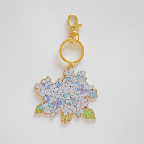The Sara Fitz Hydrangea Bloom Bag Charm is an imported gold keychain with pastel blue hydrangea flowers and green leaves in enamel stained glass style, attached to a sleek ring, shown on a plain white background.