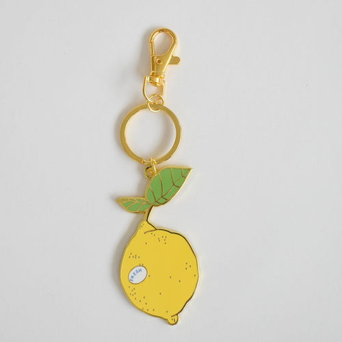 Introducing the Lemon Bag Charm by Sara Fitz—a gold keychain shaped like a yellow lemon with green leaves, adorned with a small "FRESH" sticker. This accessory features an enamel finish and a sturdy ring, and is photographed against a plain white background.