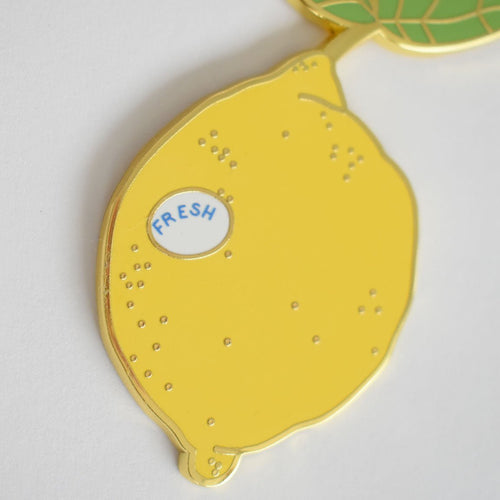 The Sara Fitz Lemon Bag Charm is a yellow enamel accessory shaped like a lemon with small dots, featuring a tiny white oval sticker printed with "FRESH" in blue letters. It is secured with a sturdy clasp and shown against a plain white background.