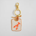 The Sara Fitz Lobster Bib Bag Charm is a gold keychain with a ring clasp, featuring a rectangular white enamel charm adorned with an orange lobster illustration and an uneven orange border, displayed on a plain white background.