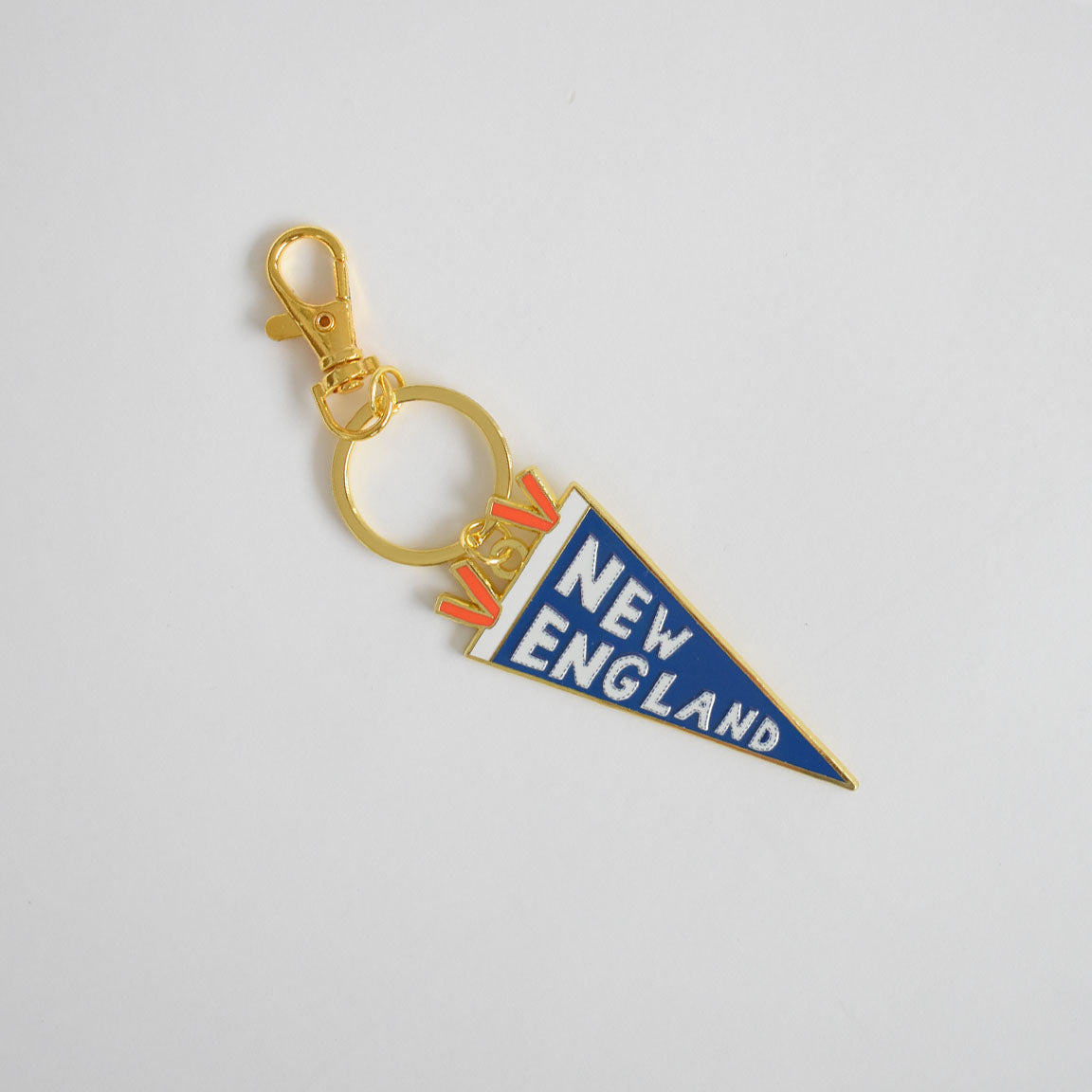 A Sara Fitz Pennant Bag Charm featuring a gold keychain with a blue enamel pennant-shaped charm that reads "NEW ENGLAND" in white letters and small red accents near the top, finished with a sturdy ring clasp and shown on a plain white background.