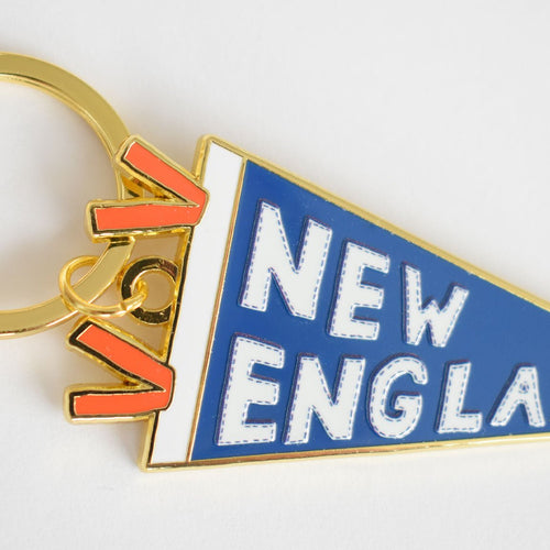 The Sara Fitz Pennant Bag Charm is a keychain shaped like a blue pennant with “NEW ENGLAND” in white letters and features a gold-colored ring clasp. Above the pennant, the word “YES!” appears in orange and gold enamel, adding a vibrant accent to this imported accessory.
