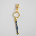 The Sara Fitz Quarterboard Bag Charm features a gold keychain with a lobster clasp and a blue vertical enamel tag displaying "SEA LA VIE" in white letters, accented by small gold seashells at the top and bottom. This stylish accessory is imported for added quality.