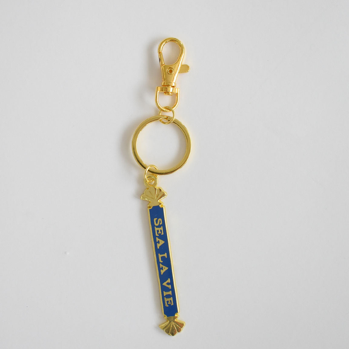 The Sara Fitz Quarterboard Bag Charm features a gold keychain with a lobster clasp and a blue vertical enamel tag displaying "SEA LA VIE" in white letters, accented by small gold seashells at the top and bottom. This stylish accessory is imported for added quality.