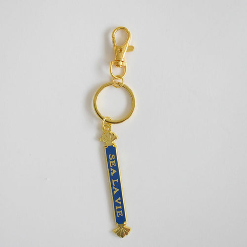 The Sara Fitz Quarterboard Bag Charm features a gold keychain with a lobster clasp and a blue vertical enamel tag displaying "SEA LA VIE" in white letters, accented by small gold seashells at the top and bottom. This stylish accessory is imported for added quality.