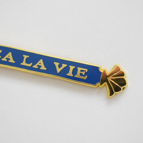 A Sara Fitz Quarterboard Bag Charm featuring gold and blue enamel with the partial inscription "LA LA VIE" and a gold shell design on the right, displayed against a white background.