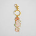 The Sara Fitz Seahorse Bag Charm features an imported enamel pink and orange seahorse, adorned with floral accents on its head and light green fins, attached to a classic gold keyring, all showcased against a plain white background.