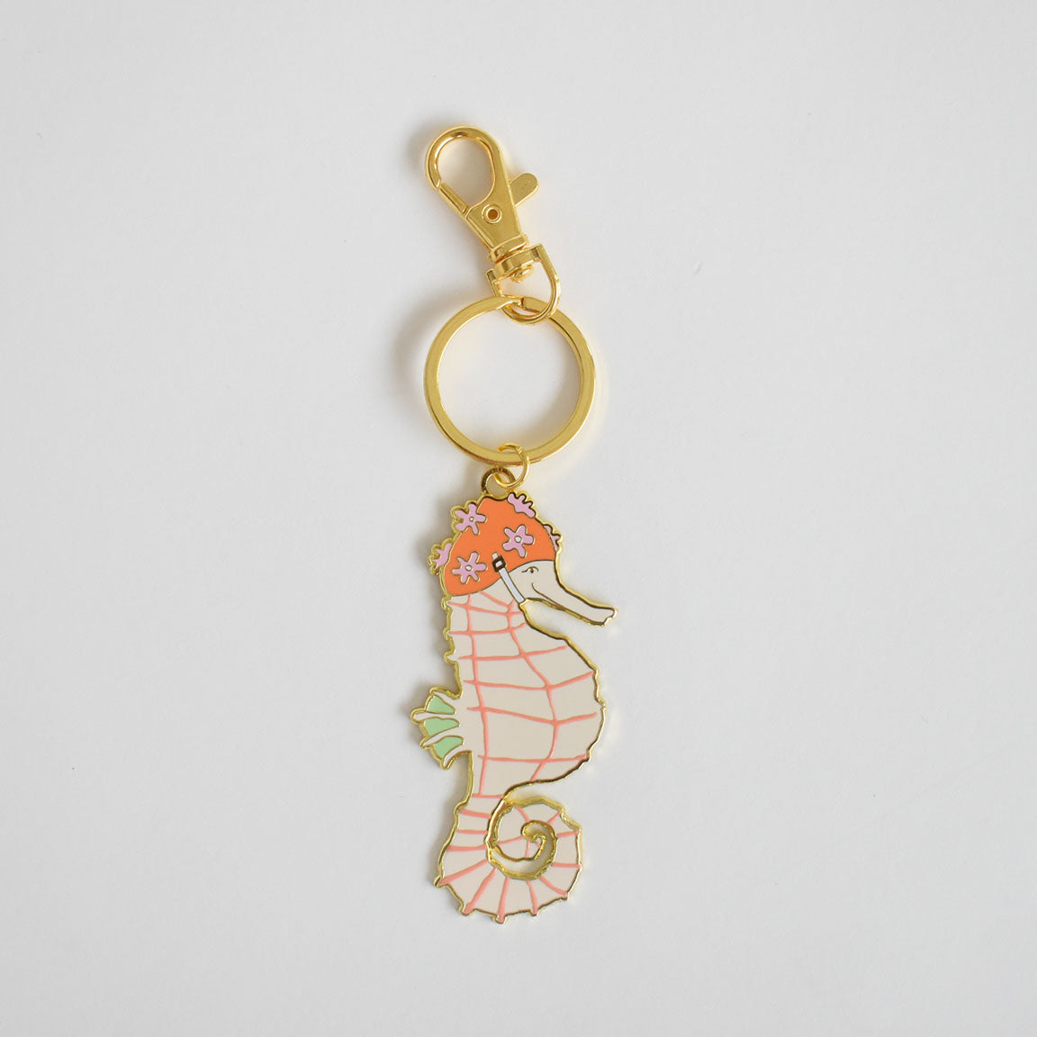 The Sara Fitz Seahorse Bag Charm features an imported enamel pink and orange seahorse, adorned with floral accents on its head and light green fins, attached to a classic gold keyring, all showcased against a plain white background.