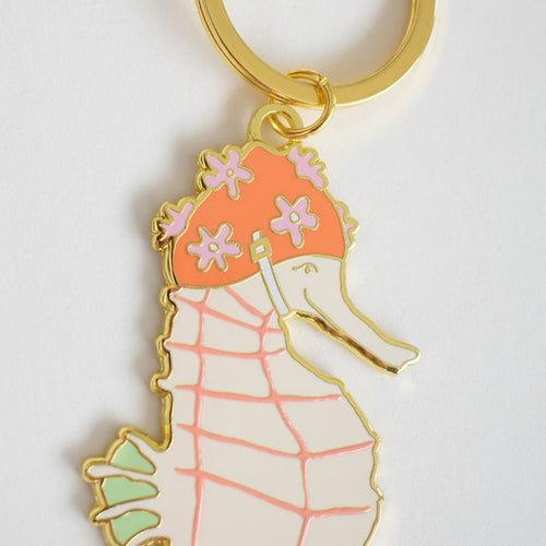 Sara Fitz Seahorse Bag Charm: An imported gold keychain featuring a pastel enamel seahorse wearing a pink flower-adorned shower cap, attached to a sturdy ring.