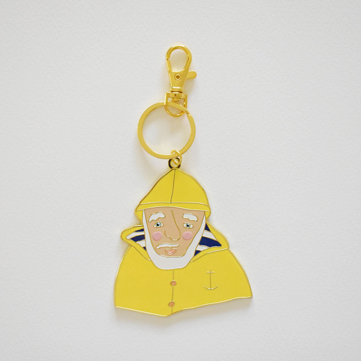 The Skipper Bag Charm by Sara Fitz is an imported keychain featuring a cartoon-style figure of an older person in a yellow enamel raincoat and hat, accented with a gold anchor symbol, set against a plain white background.