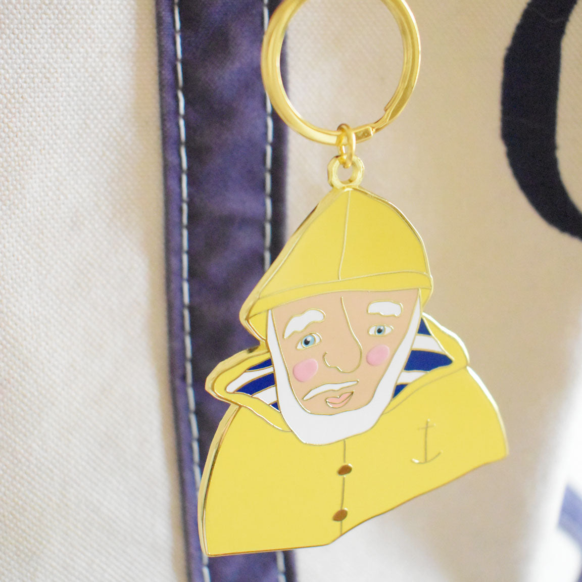 The Skipper Bag Charm by Sara Fitz is an imported enamel keychain that features an illustration of a bearded man in a yellow raincoat and hat, with pink cheeks, a blue striped shirt, and an anchor detail, hanging on a bag with white and navy fabric.