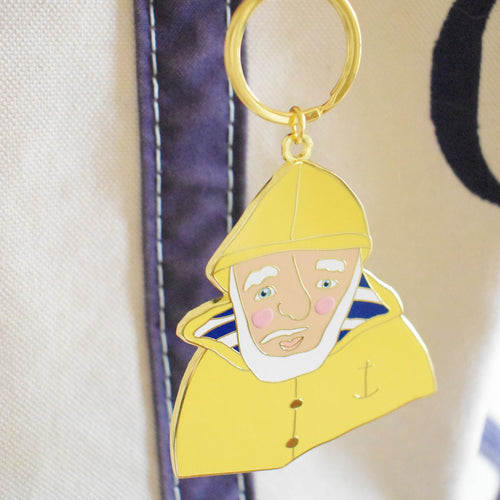 The Skipper Bag Charm by Sara Fitz is an imported enamel keychain that features an illustration of a bearded man in a yellow raincoat and hat, with pink cheeks, a blue striped shirt, and an anchor detail, hanging on a bag with white and navy fabric.