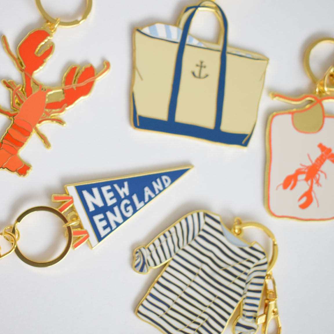 Five imported Sara Fitz enamel Pennant Bag Charms—a red lobster, a tan tote bag with an anchor, a lobster bib, a blue-striped shirt, and a blue pennant reading "New England," all on sturdy ring clasps—are set against a white background.